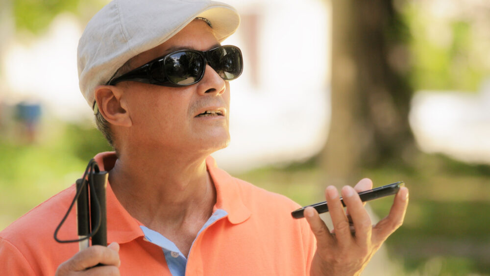 A visually impaired man with sunglasses and a hat holding a cell phone
