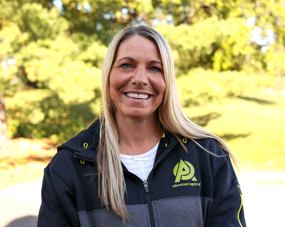 A cheerful Cheryl Borger, an optical frames lab technician from Allentown, wearing a black and yellow jacket, smiles brightly.